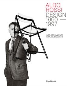 Book cover of Aldo Rossi: Design - 1960-1997. Catalogue Raisonné, with the designer grasping a black framed clear Perspex chair in left hand. Published by Silvana.