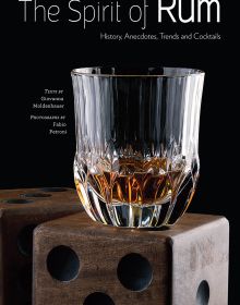 Book cover of The Spirit of Rum, History, Anecdotes, Trends and Cocktails, with a crystal glass of rum on large wooden die. Published by White Star.