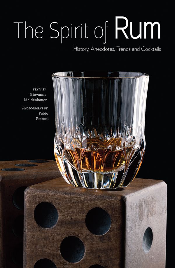 Book cover of The Spirit of Rum, History, Anecdotes, Trends and Cocktails, with a crystal glass of rum on large wooden die. Published by White Star.