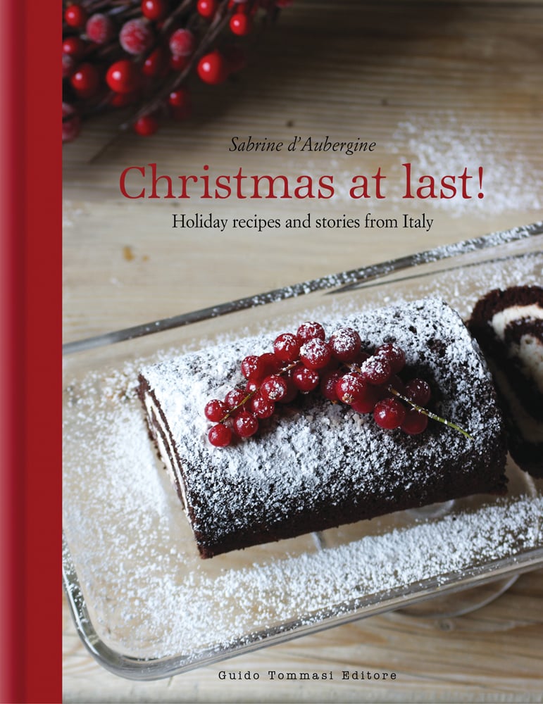 Christmas chocolate roulade, sprinkled with icing sugar, on glass dish, on cover of 'Christmas at Last! Holiday Recipes and Stories from Italy', by Guido Tommasi Editore.