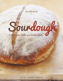 Large round brown top sourdough bread, dusted with flour, on wood board, on cover of 'Sourdough, A Complete Guide and Recipe Book', by Guido Tommasi Editore.