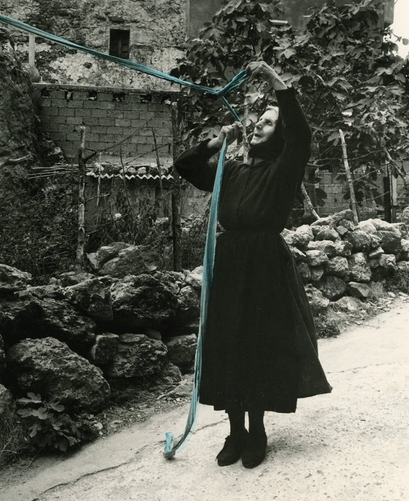 Woman dressed all in black stretching out a long blue ribbon, in town setting with rocks edging roadside