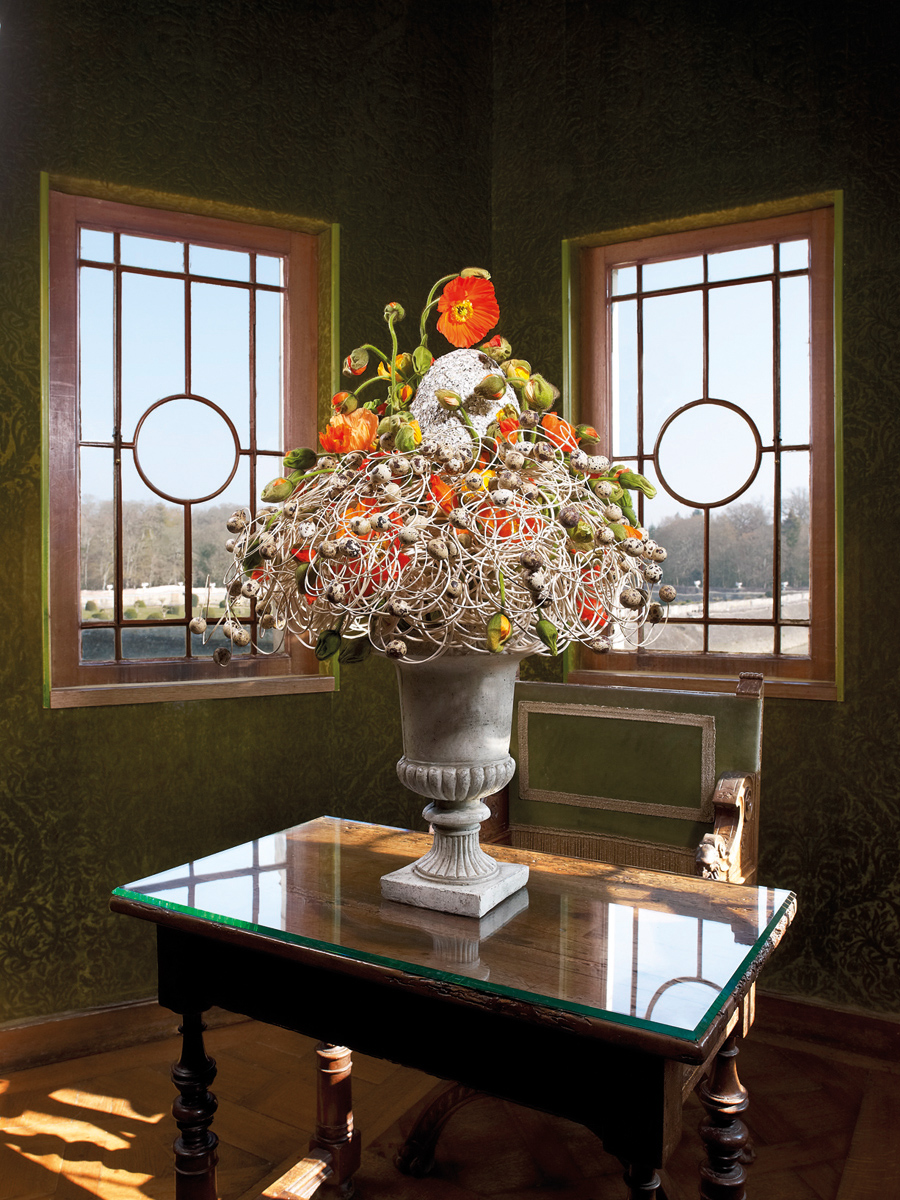 Bouquet of flowers in urn with base, in front of decorative fireplace, THE BOUQUETS OF CHENONCEAU in bright yellow font above.