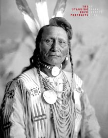 Black and white portrait of Fort Yate Native American tribesman, feather in hair, beads around neck, on cover of 'The Standing Rock Portraits, Sioux Photographed by Frank Bennett Fiske 1900-1915', by Lannoo Publishers.