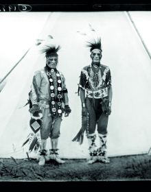 Black and white portrait of Fort Yate Native American tribesman, feather in hair, beads around neck, on cover of 'The Standing Rock Portraits, Sioux Photographed by Frank Bennett Fiske 1900-1915', by Lannoo Publishers.