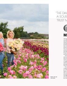 Figure in pink wellington boots, holding cream bucket filled with pink dahlias, surrounded by field of flowers, on cover of 'The Joy of Dahlias', by Lannoo Publishers.
