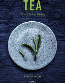 Green tea leaves on pale blue plate, deep blue tablecloth beneath, on cover of 'Tea', Wine's Sober Sibling', by Lannoo Publishers.