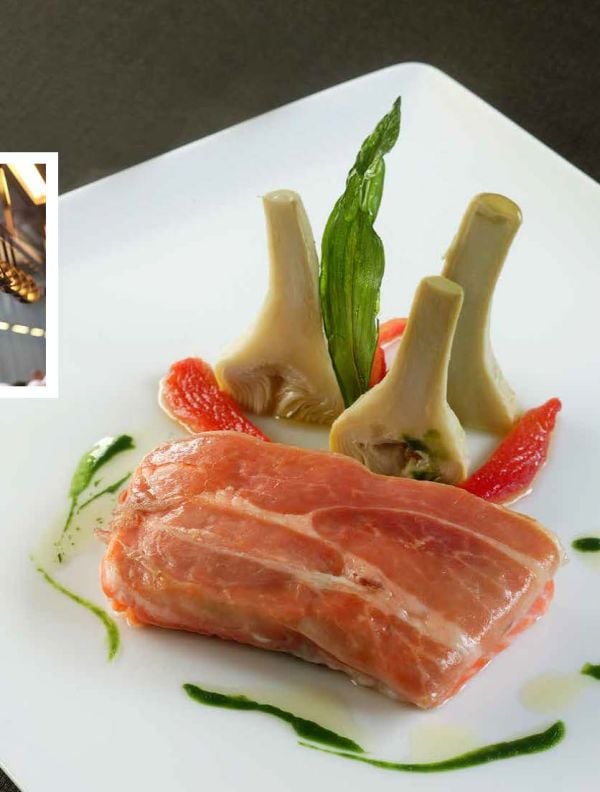 Ham hock with metal hook, tied with string, on charcoal cover of 'Ham, Prime Hams of Europe Stories and Recipes', by Lannoo Publishers.