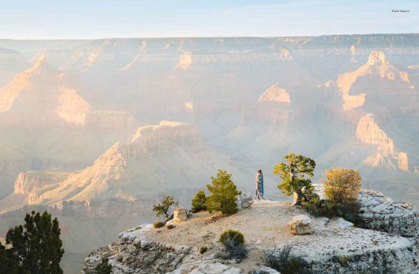 Campervan on straight road with mountainous landscape behind and orange sky, on cover of 'Roaming America, Exploring All the National Parks', by Lannoo Publishers.