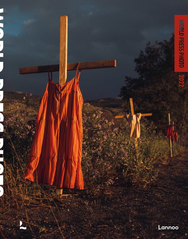 Book cover of World Press Photo 2022, with orange and cream summer dresses hanging from three wooden crosses in ground. Published by Lannoo Publishers.