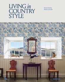 Interior with white panelled walls, blue floral wallpaper above, two windows looking out to hilly landscape, on cover of 'Living in Country Style', by Lannoo Publishers.