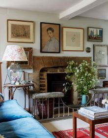 Interior with white panelled walls, blue floral wallpaper above, two windows looking out to hilly landscape, on cover of 'Living in Country Style', by Lannoo Publishers.