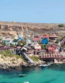 Row of multicolored slated wood chairs, near harbour edge, with boat, on cover of Hidden Malta', by Luster Publishing.