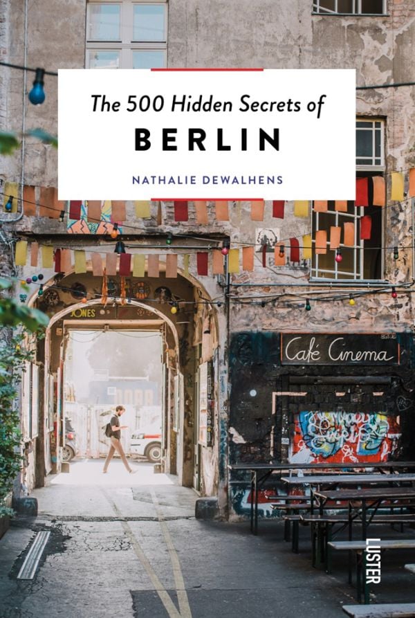 Book cover of The 500 Hidden Secrets of Berlin, with Café Cinema bench with bunting and street art. Published by Luster Publishing.