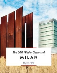 Tall rusted metal panelled structure surrounded by pale dry grass, tall building behind, The 500 Hidden Secrets of MILAN in black font on bottom white banner.