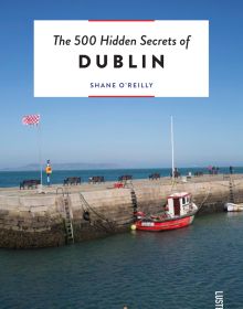 Book cover of The 500 Hidden Secrets of Dublin, with a fishing boat at Bulloch Harbour, Dalkey, under blue sky. Published by Luster Publishing.