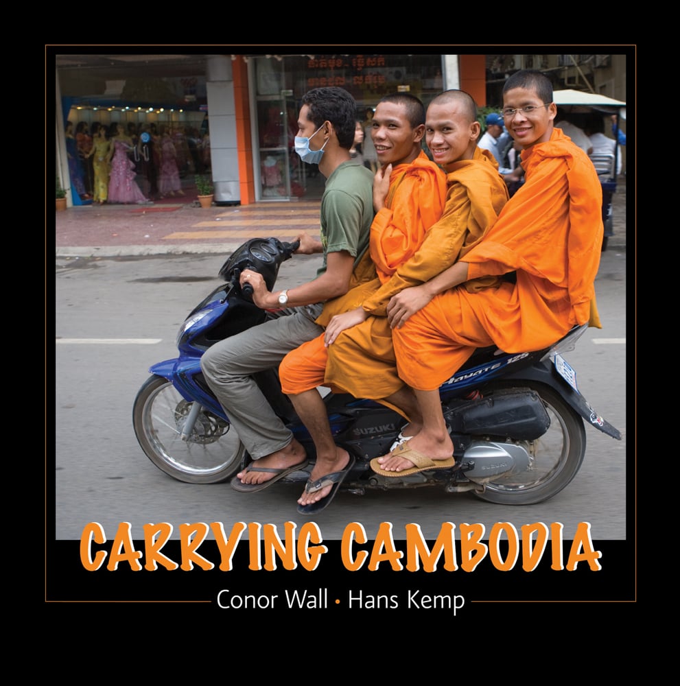 Vietnamese biker with 3 novice Buddhist monks in orange robes on back, riding through street, black border, CARRYING CAMBODIA in orange font with white drop shadow, below.