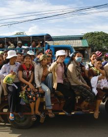 Vietnamese biker with 3 novice Buddhist monks in orange robes on back, riding through street, black border, CARRYING CAMBODIA in orange font with white drop shadow, below.