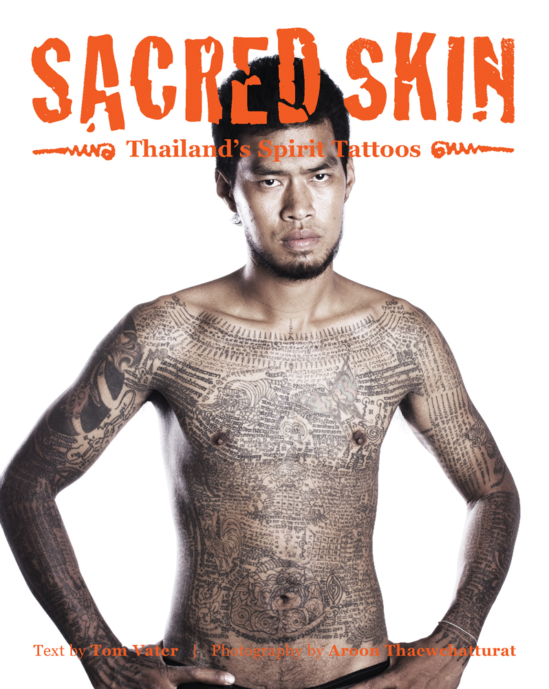 Asian male, hands on hips, serious face, with torso covered in finely detailed black tattoos, SACRED SKIN Thailand's Spirit Tattoos in orange font above.