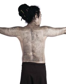 Asian male, hands on hips, serious face, with torso covered in finely detailed black tattoos, SACRED SKIN Thailand's Spirit Tattoos in orange font above.