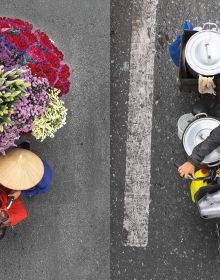 Aerial view of 2 Vietnamese street vendors in leaf hats, next to bikes with trailers of colourful fruits, MERCHANTS IN MOTION in white font above.