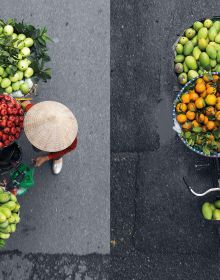 Aerial view of 2 Vietnamese street vendors in leaf hats, next to bikes with trailers of colourful fruits, MERCHANTS IN MOTION in white font above.